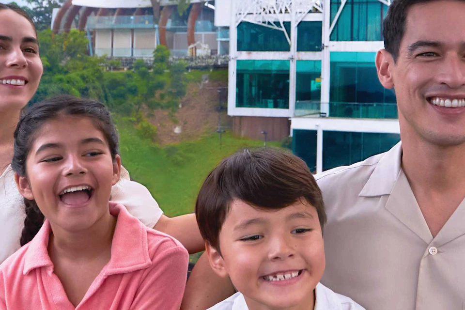 A family of four smiles inside a cable car with a scenic view of greenery and buildings outside.