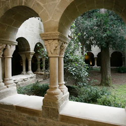 Stenbågar och detaljerade pelare i ett kloster omger en frodig innergård med träd och grönska.