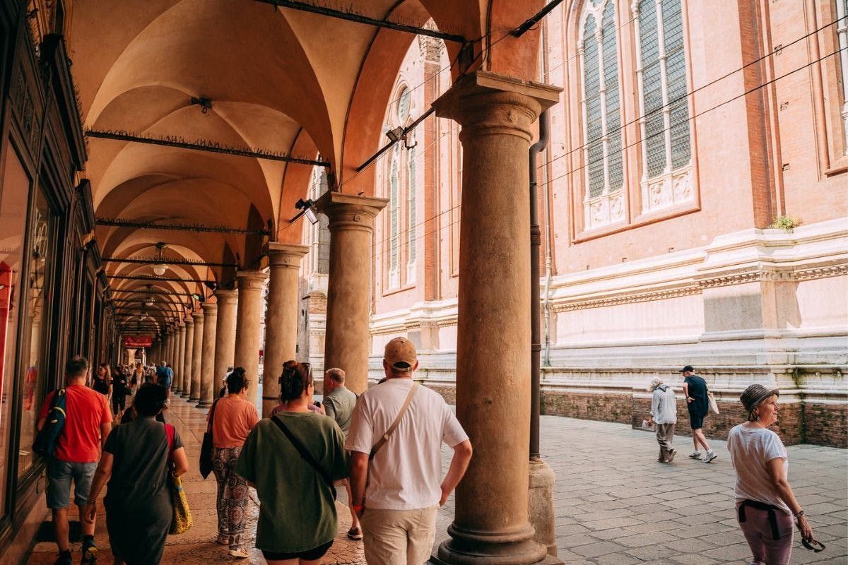 People walking under an arched colonnade with tall columns next to a historical brick building with large windows.