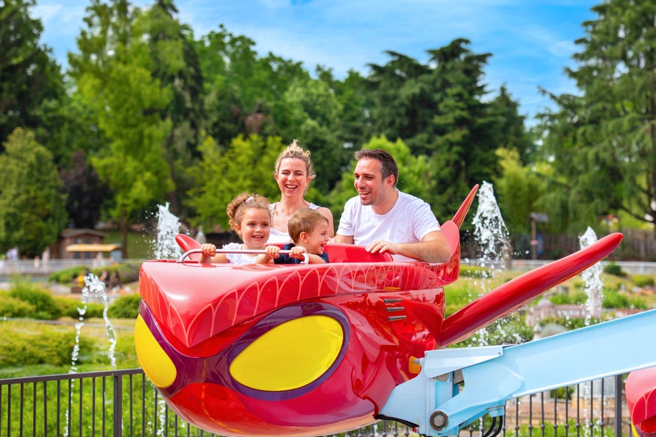 A family of four enjoys a colorful amusement park ride shaped like a cartoon bird, with fountains and trees in the background.