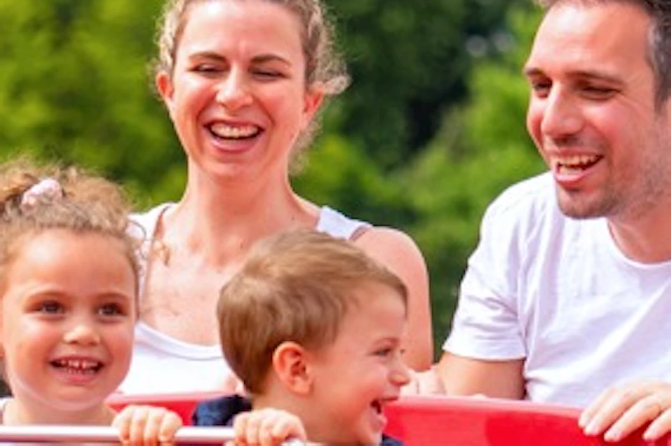 A family of four enjoys a colourful amusement park ride shaped like a cartoon bird, with fountains and trees in the background.
