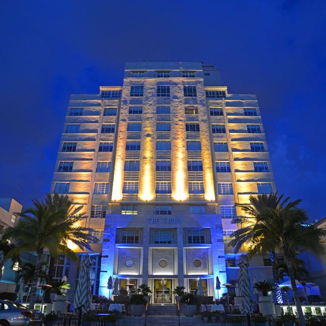 Tall building illuminated at night with warm lights, flanked by palm trees, set against a deep blue sky.