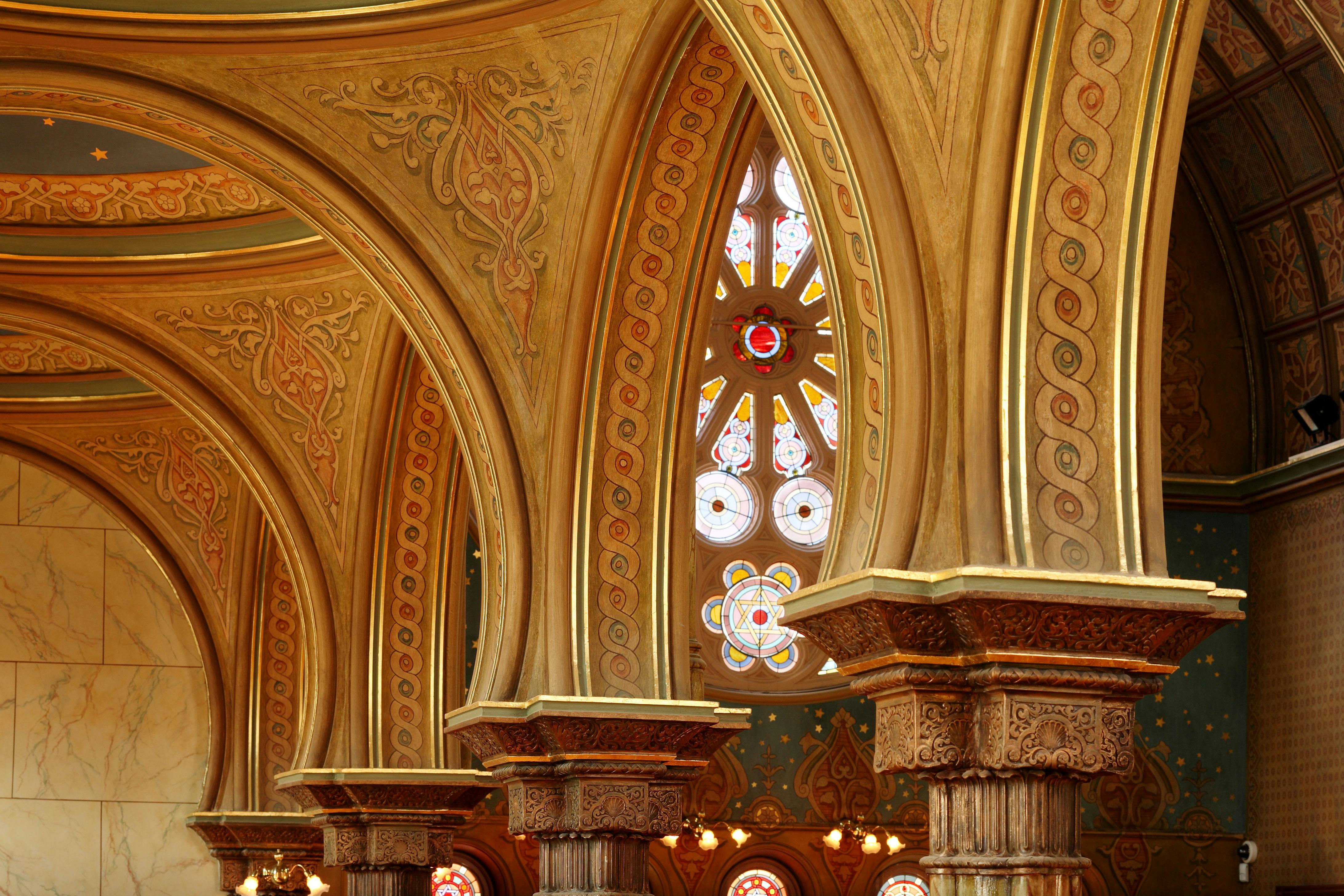 Des arcades et des colonnes ornées de motifs complexes et un vitrail aux motifs géométriques dans un intérieur richement décoré.
