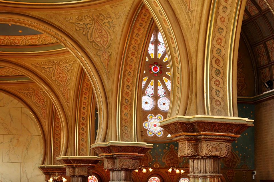 Ornate archways and columns with intricate designs and a stained glass window featuring geometric patterns in a richly adorned interior.