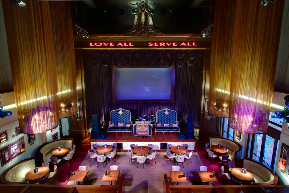 A stylish lounge with round tables and velvet chairs, a large screen, and illuminated signs reading "LOVE ALL" and "SERVE ALL."
