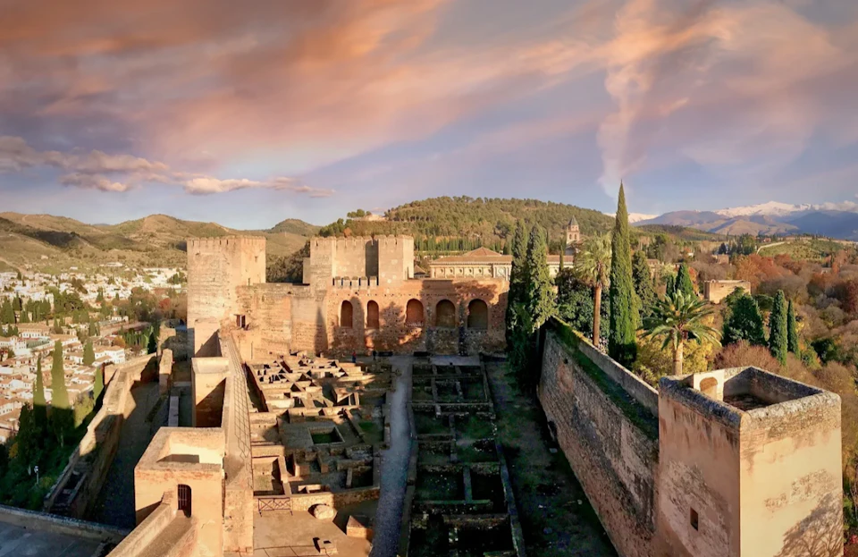 A historic fortress with stone walls, towers, and ruins, surrounded by greenery and a scenic landscape under a colorful sky.