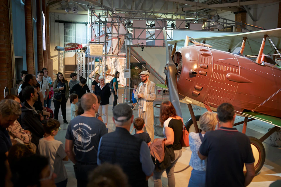 A group of people listening to a presenter in front of a vintage airplane in an indoor museum setting.