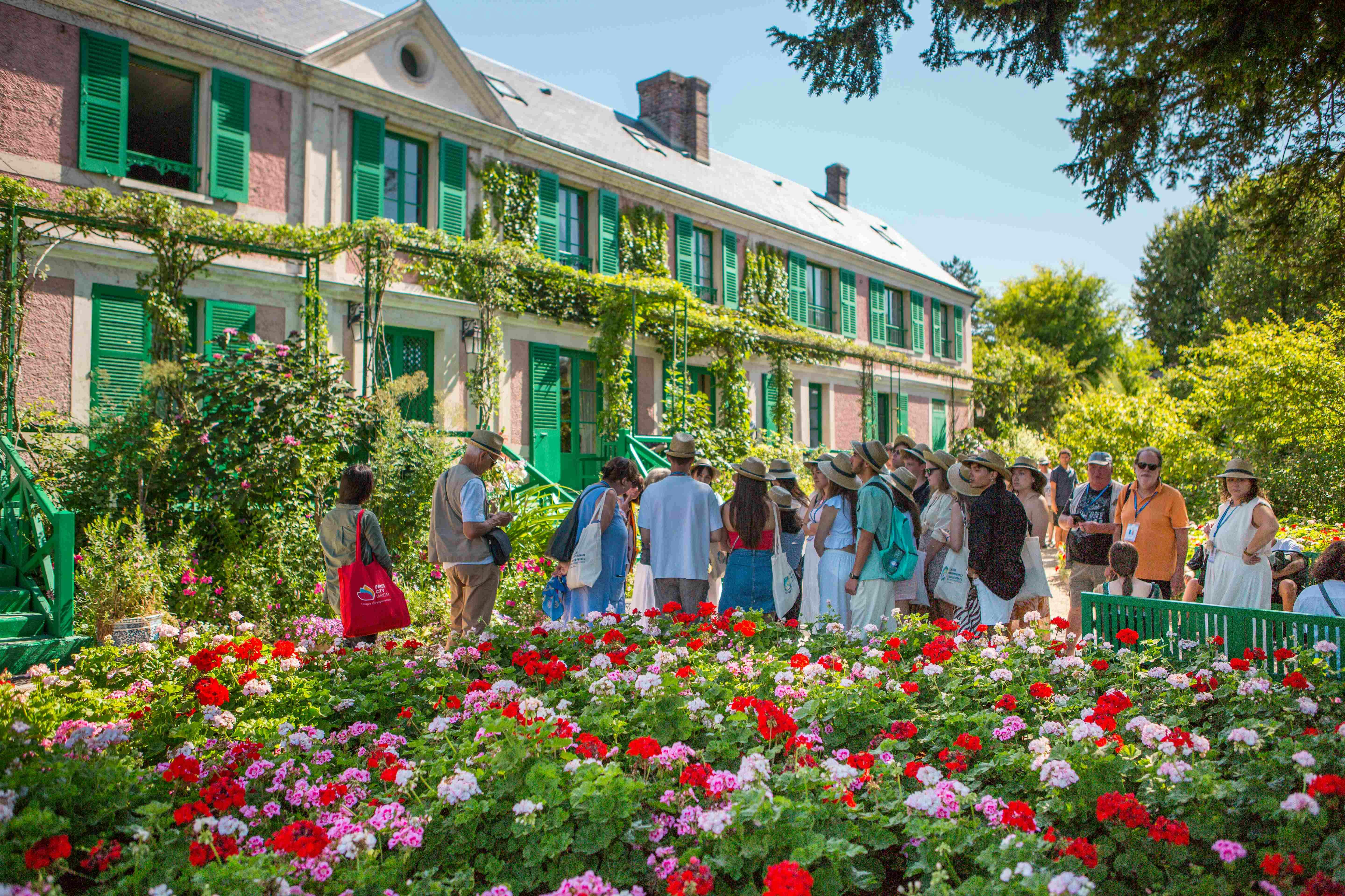 A group of people, some wearing hats, gather in a colorful flower garden in front of a vine-covered building with green shutters.