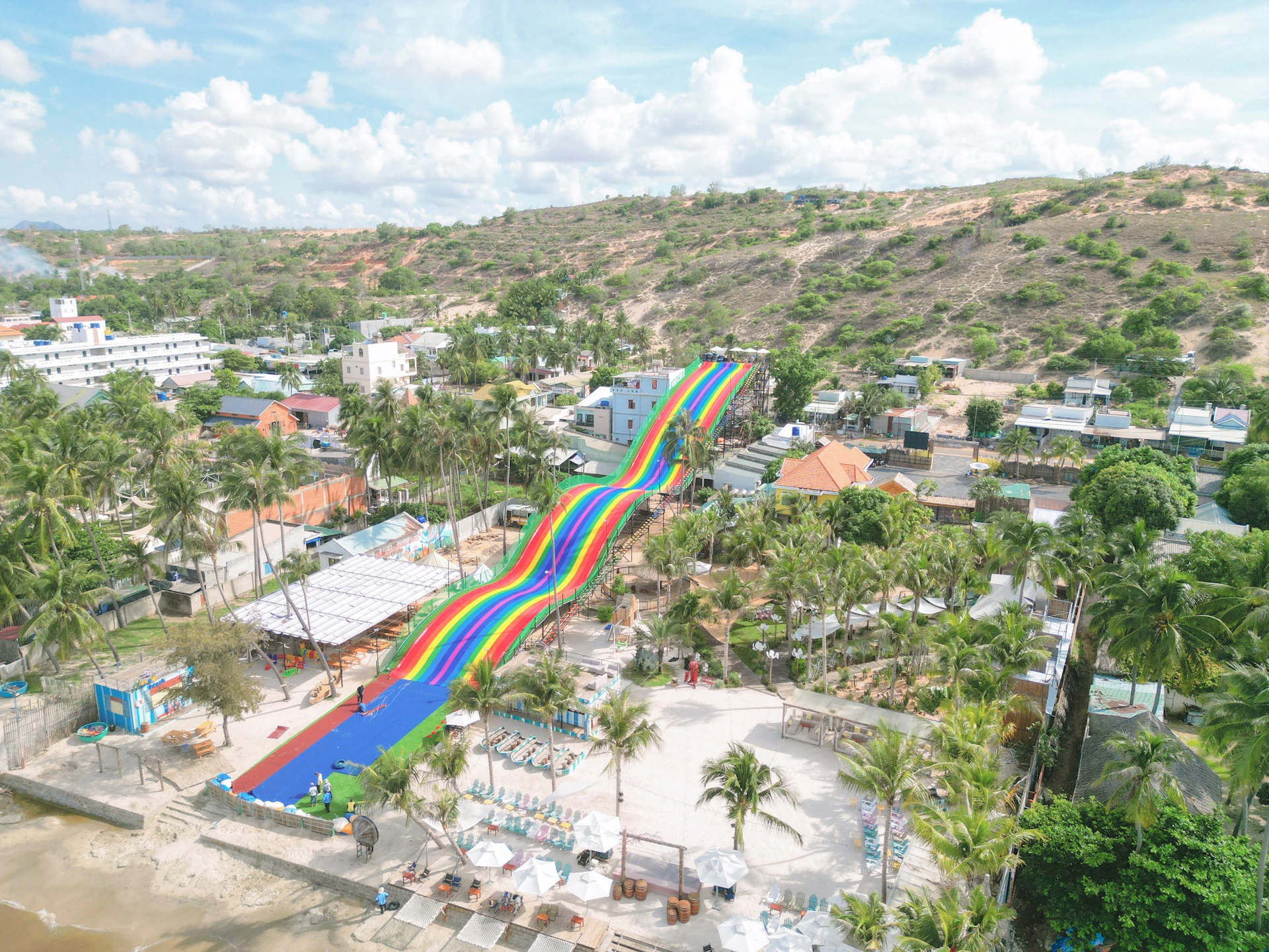 A colorful, rainbow-themed water slide stretching down a hill in a tropical setting with palm trees and buildings nearby.