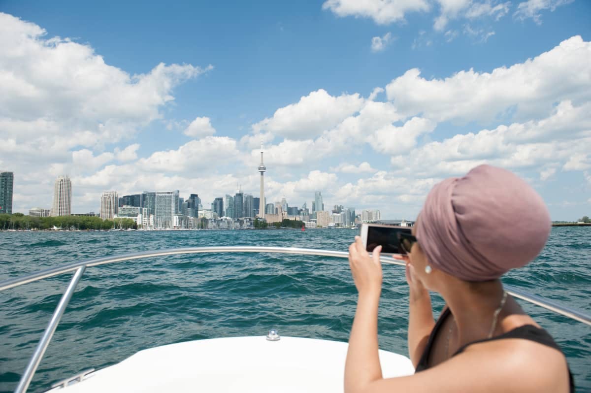 Person on a boat photographing a city skyline with a prominent tower, under a bright blue sky with scattered clouds.