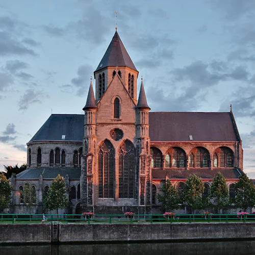 En historisk kyrka med välvda fönster och dubbla spiror, badade i solnedgångsljus, står nära en flod med en linje av träd framför.