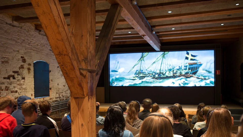 People seated in a wooden-beamed room, watching a large screen showing a sailing ship on rough seas.