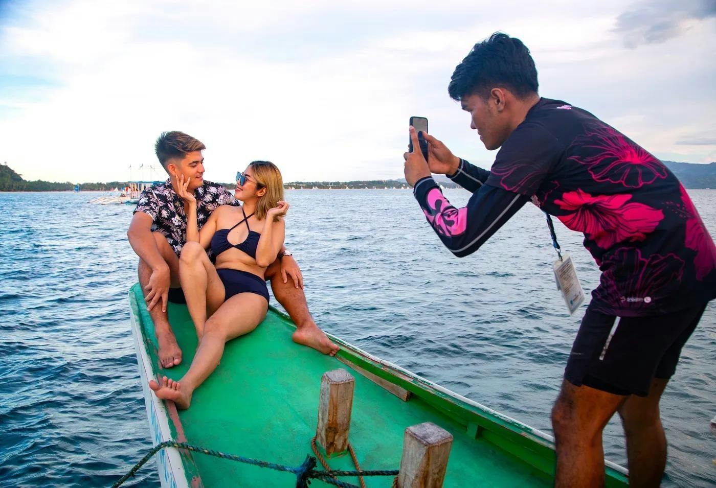 A couple poses on a small boat with a man taking their photo using a smartphone, set against a body of water and sky.