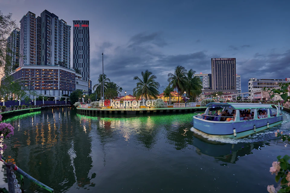 Waterfront area at dusk with palm trees, buildings, and neon-lit "Kampung Morten" sign. A boat on the river in the foreground.