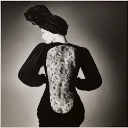 En kvinna i en mörk klänning med en ryggpanel i spets, bär huvudbindel, poserar med ryggen vänd och huvudet lutat åt sidan.