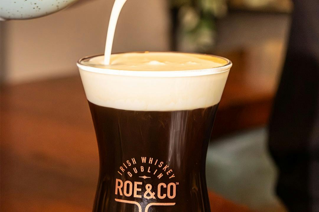 A glass of Irish coffee with cream being poured on top, branded with 'Roe & Co' and 'Irish Whiskey Dublin' text.