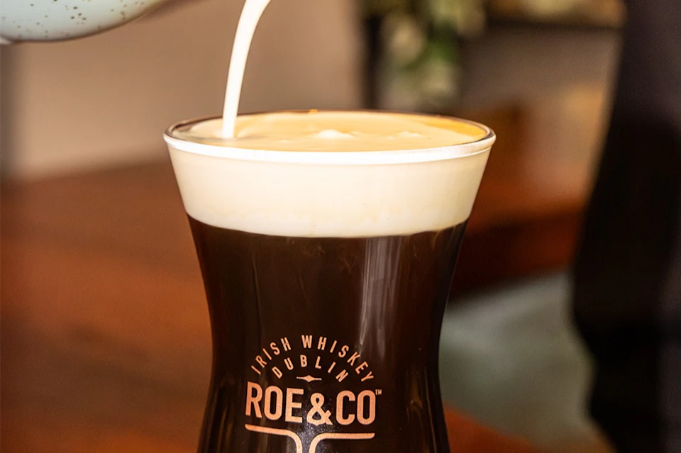 A glass of Irish coffee with cream being poured on top, branded with 'Roe & Co' and 'Irish Whiskey Dublin' text.