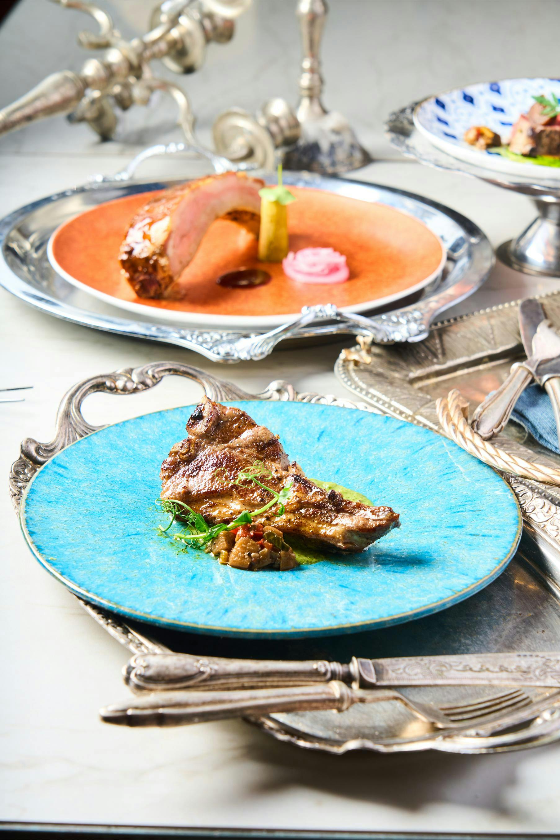 A gourmet meal featuring grilled meat on a blue plate, garnished with greens, set beside a second plate on a white table.