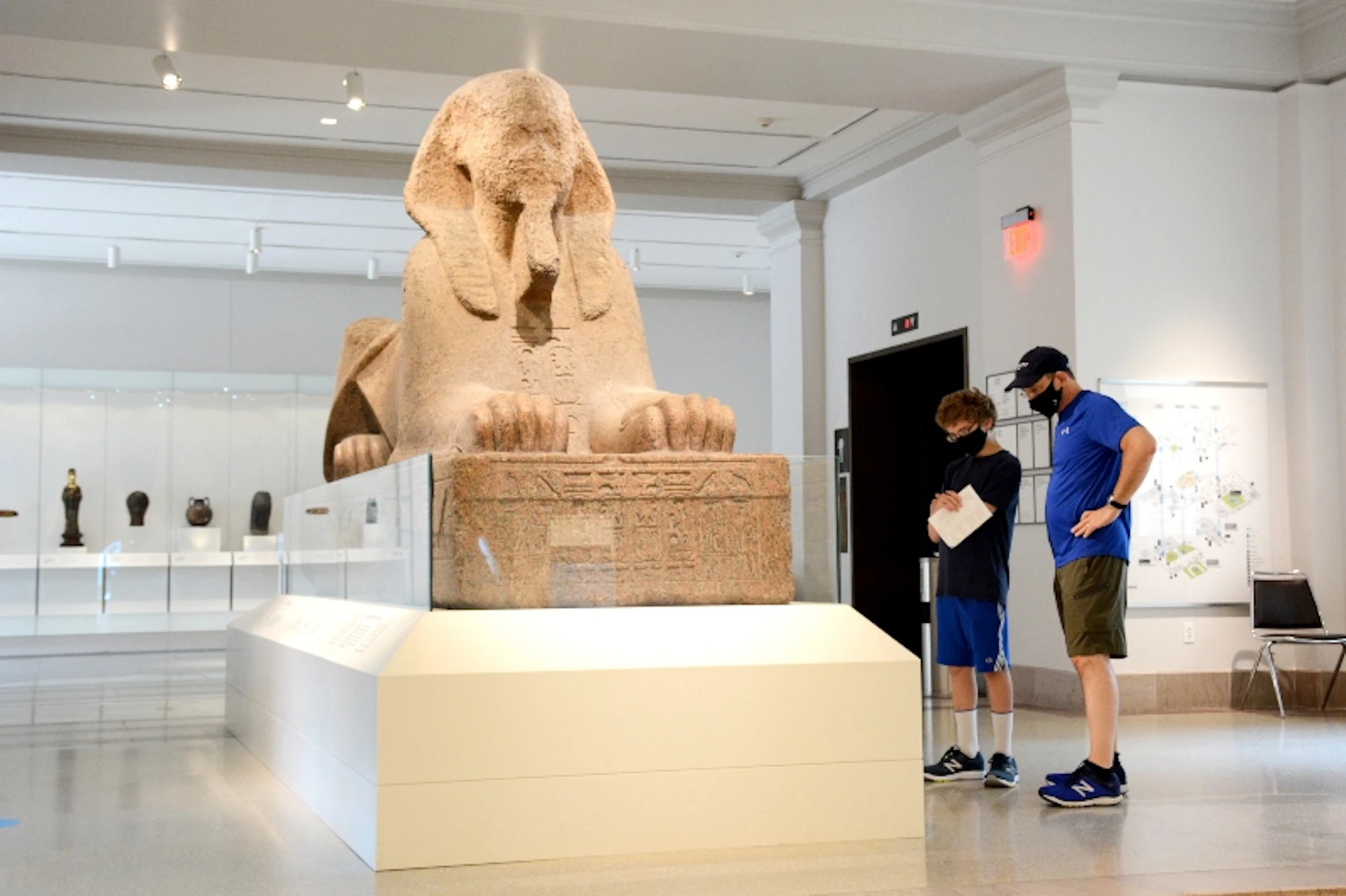 Two people standing in a museum observing a large sphinx statue encased in glass.