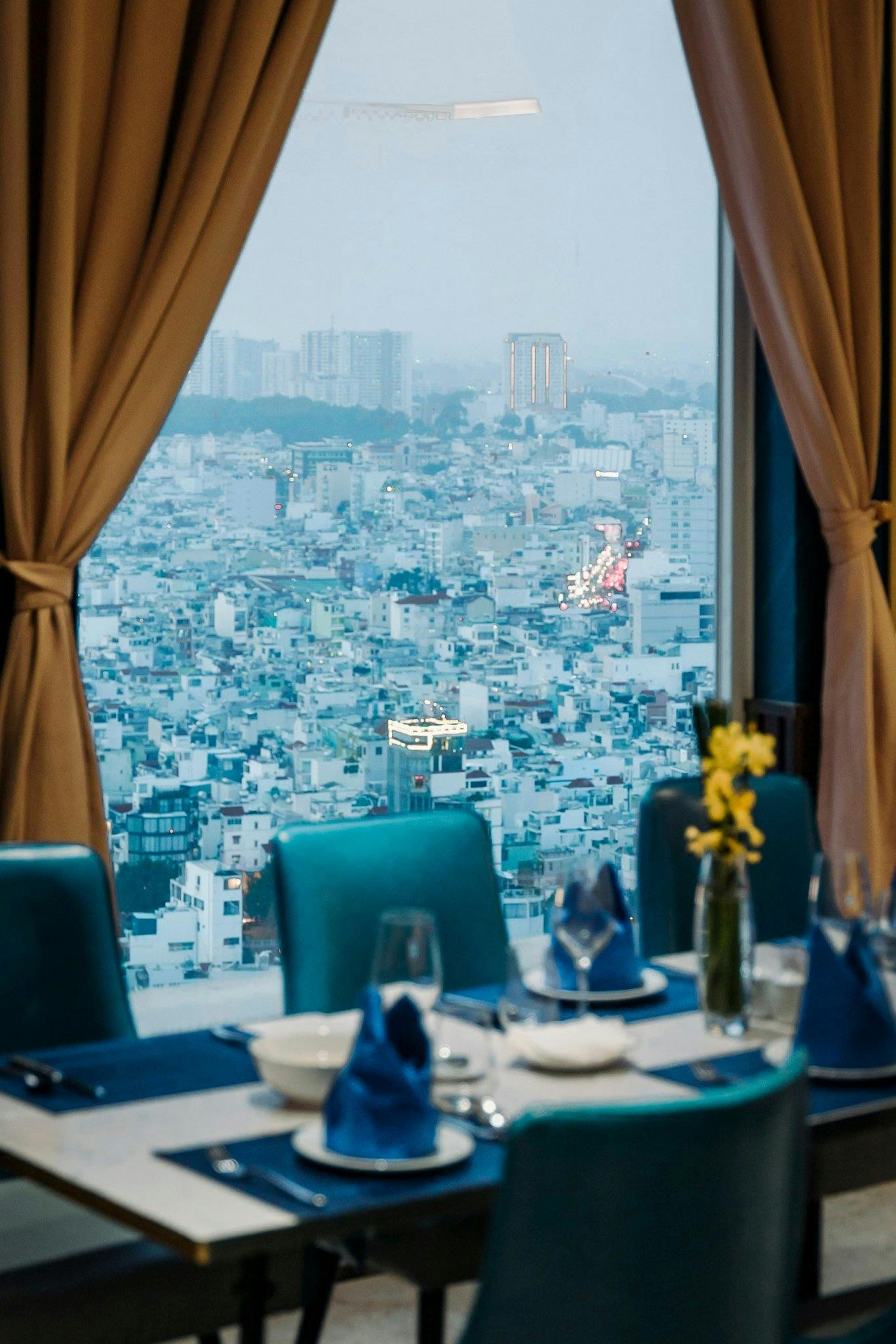 A view of a cityscape through a window with brown curtains, showing a dining table set with plates and glasses in the foreground.