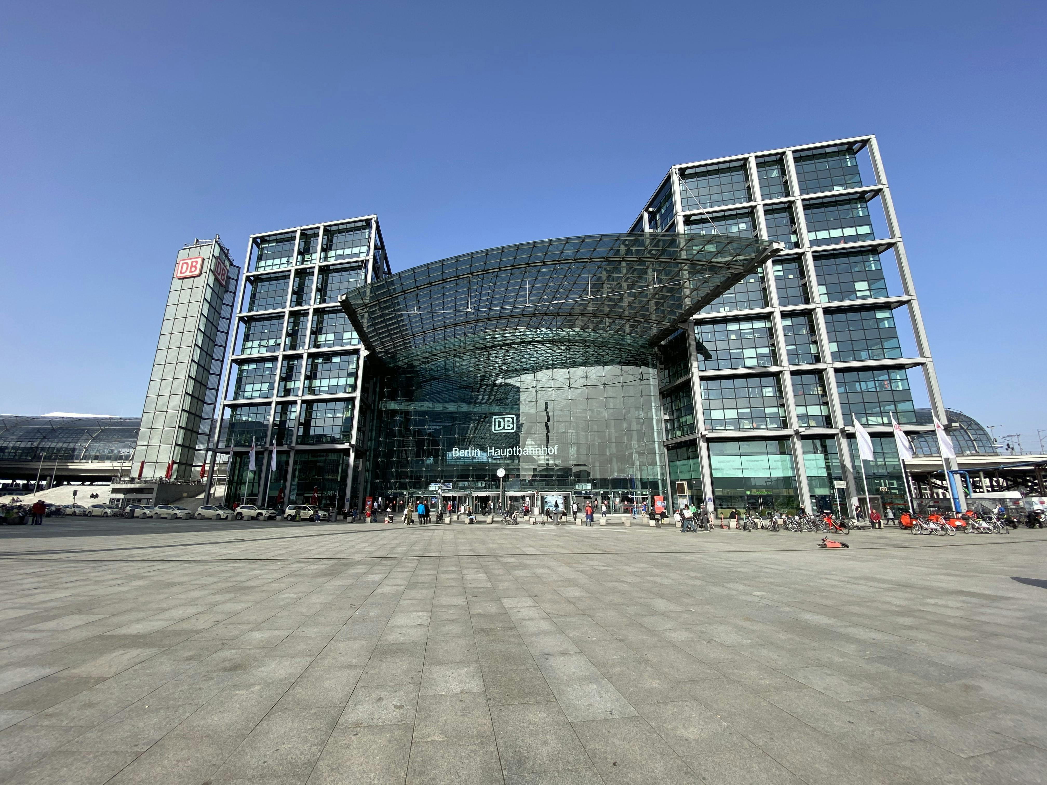 맑고 푸른 하늘 아래 커다란 석조 광장에 사람들이 모여 있는 베를린 중앙역의 현대적인 유리 건물 입구.