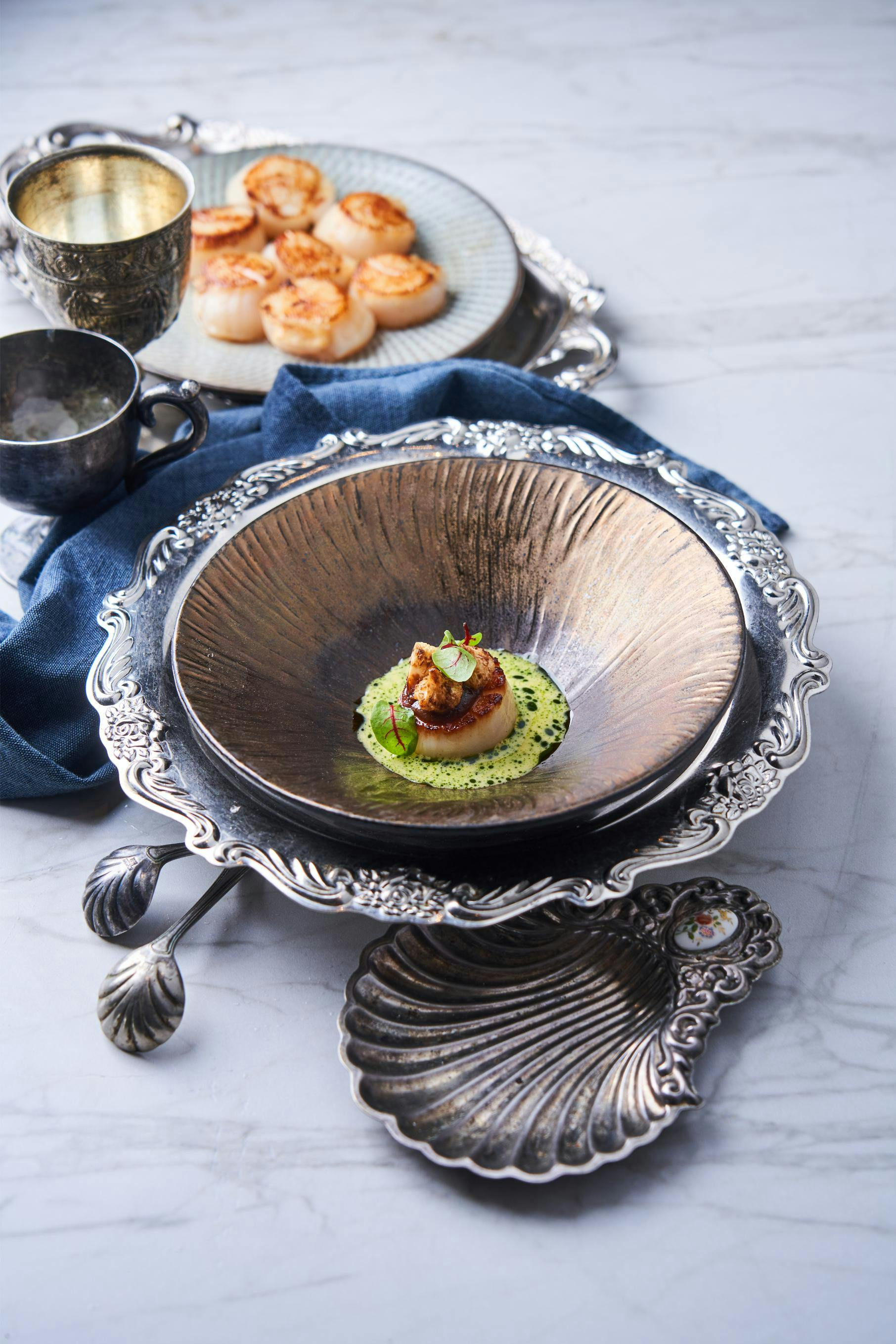 A plated scallop dish garnished with herbs in a decorative metal bowl, surrounded by ornate tableware on a marble surface.