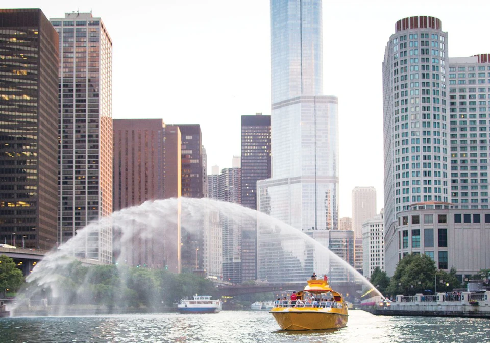 A yellow boat cruises on a river in a city, with skyscrapers in the background and water jets arching over the river.