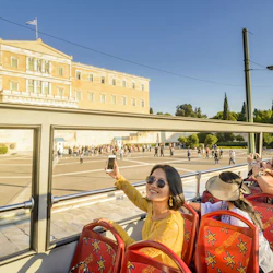 En kvinna i solglasögon tar en selfie på en turistbuss med passagerare, framför en stor historisk byggnad under en klarblå himmel.