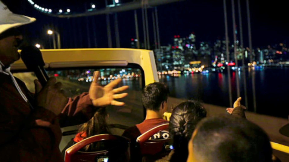 People on an open-top bus at night with a city skyline and bridge in the background.
