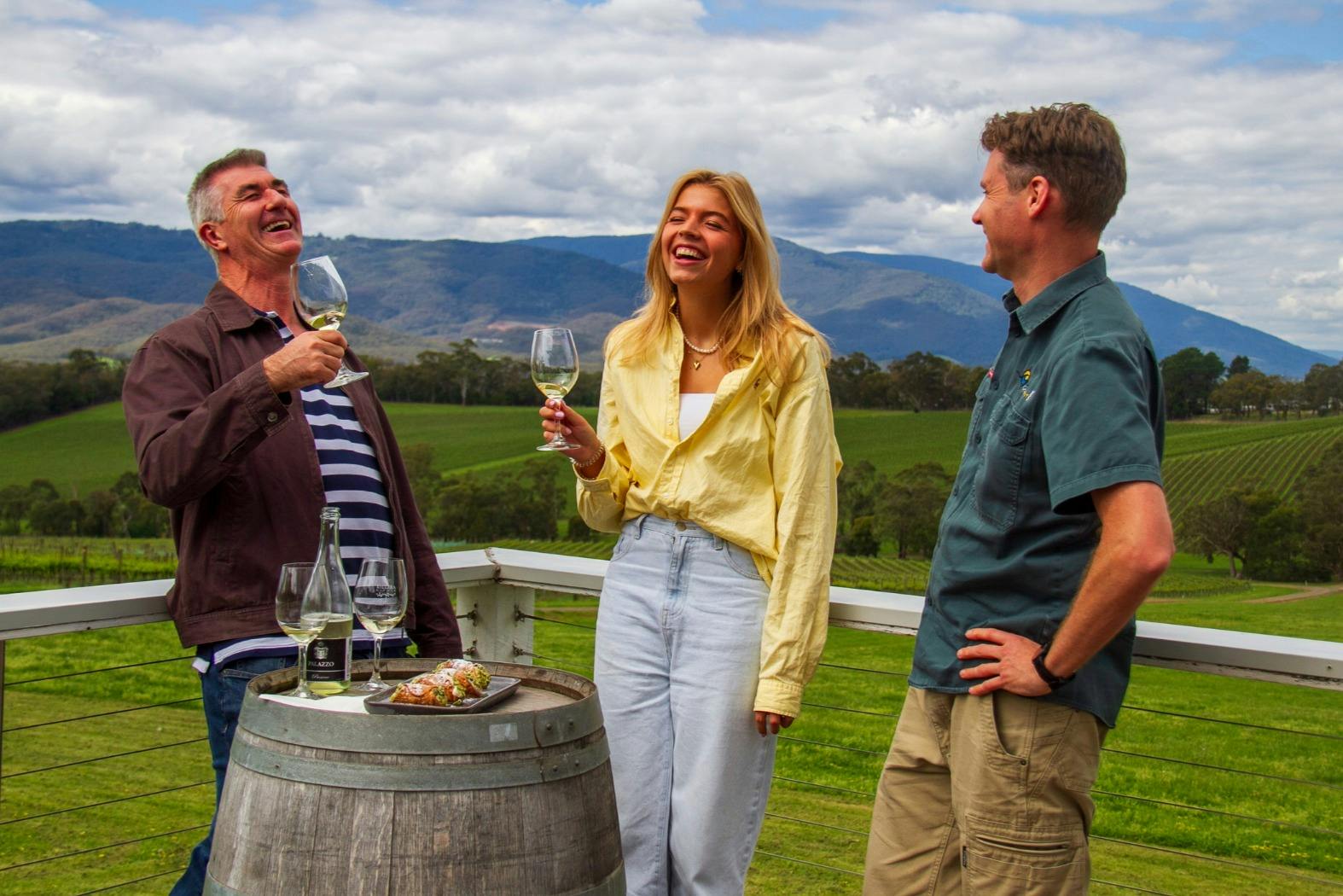 Three people standing outdoors, holding wine glasses, near a wine barrel with a picturesque vineyard in the background.
