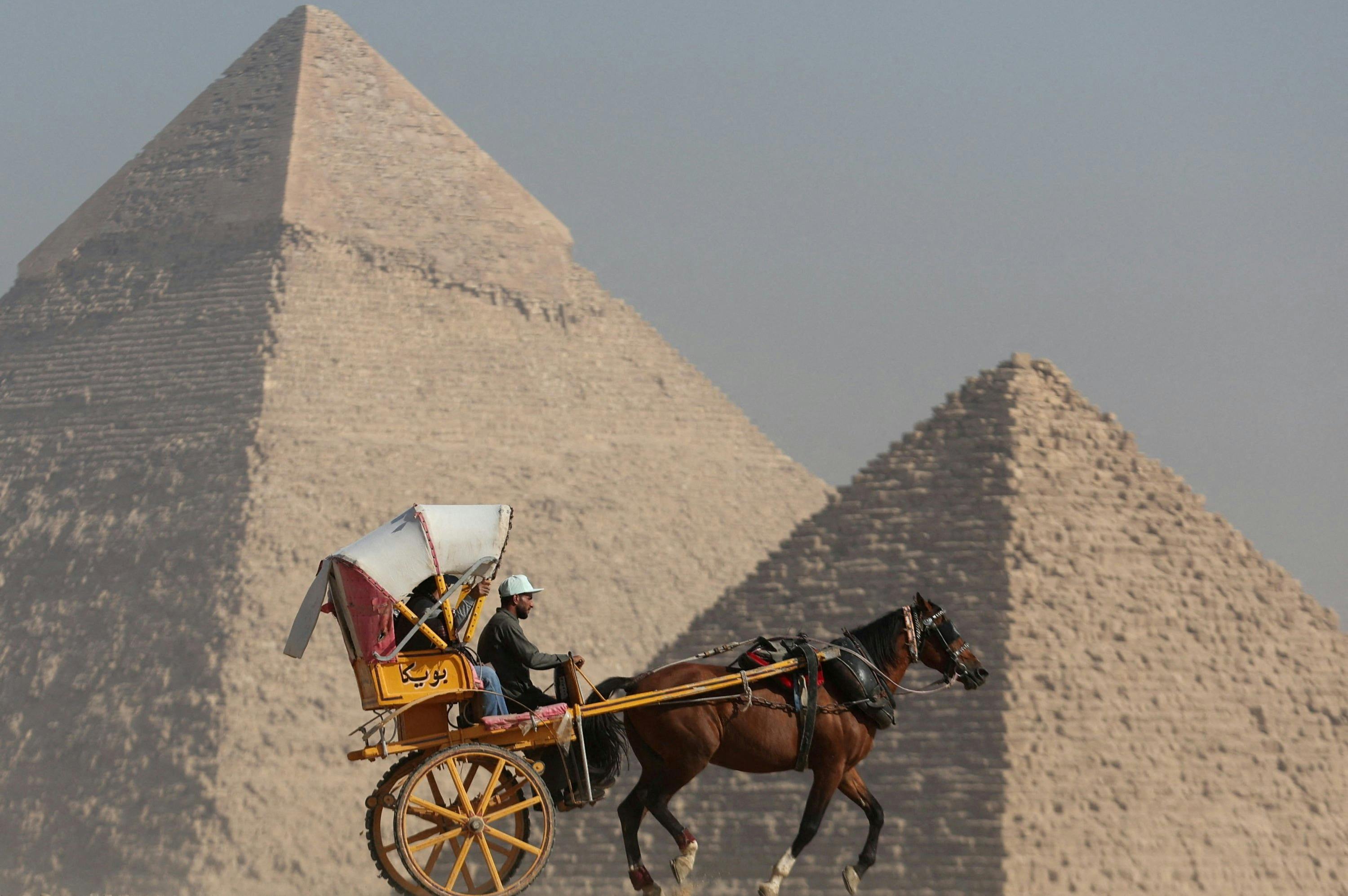 A person riding a horse-drawn carriage in front of the Pyramids of Giza under a clear sky.