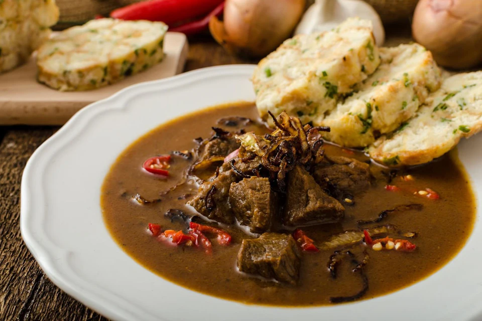 A white plate with beef stew garnished with fried onions and red peppers, served with slices of bread dumplings.