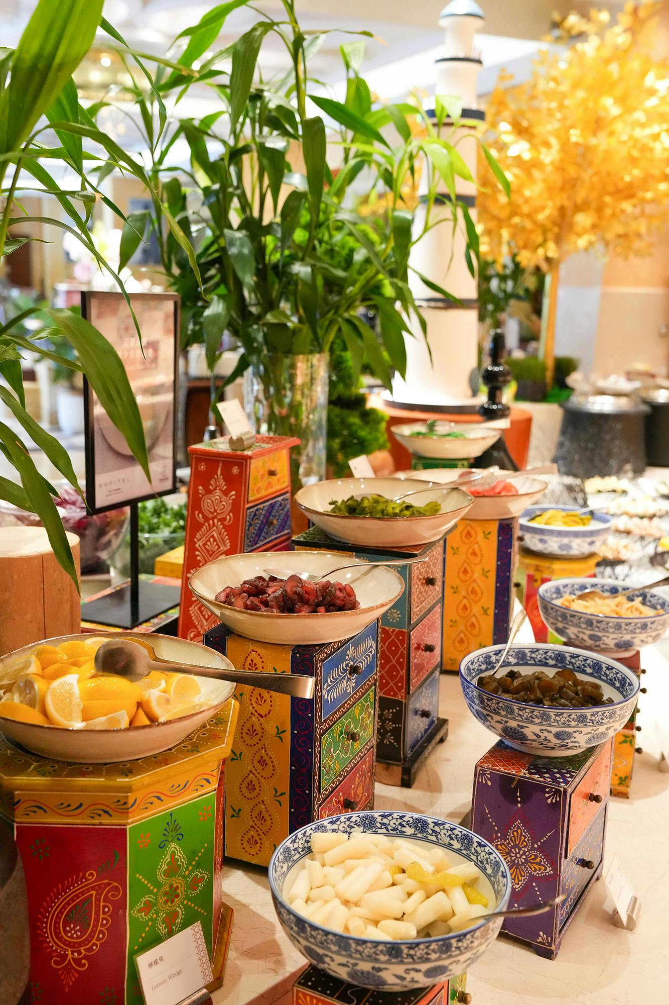 A vibrant buffet setup with colorful bowls on decorative stands, displaying various fresh ingredients and garnishes.