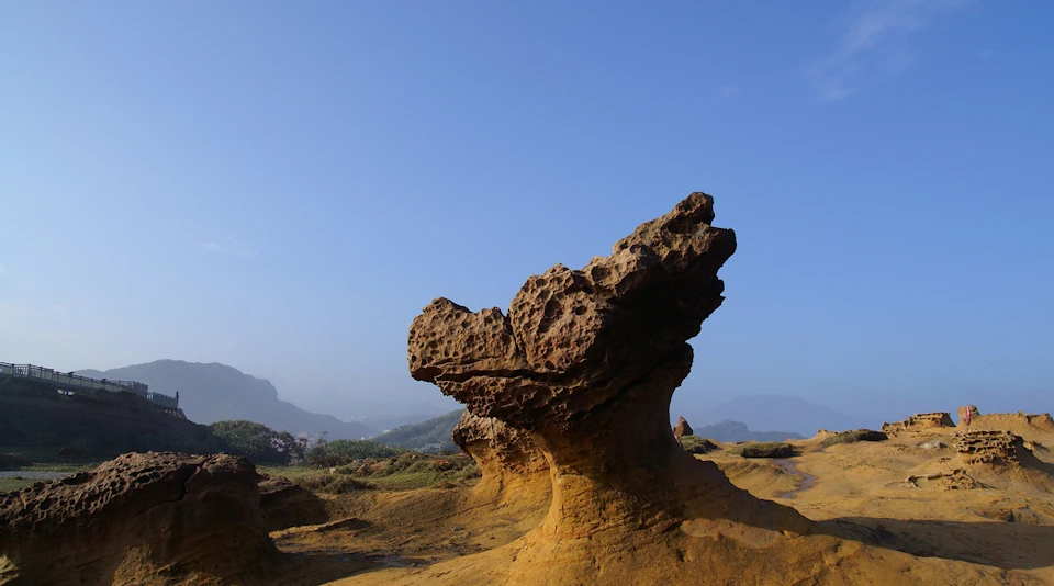 A uniquely shaped rock formation in a sunny landscape with a blue sky and distant hills.