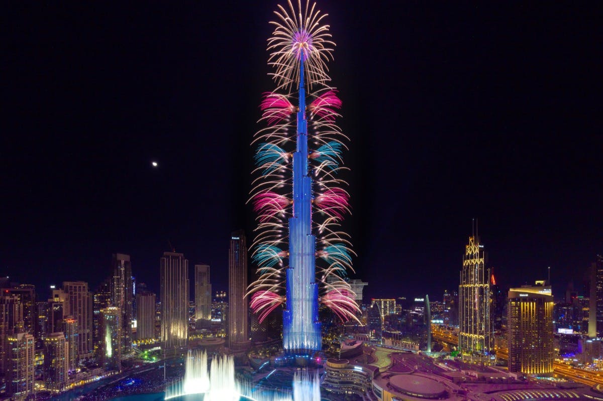 夜晚，在城市景观中，烟花照亮了一座高耸的摩天大楼，下面还有其他建筑和喷泉。