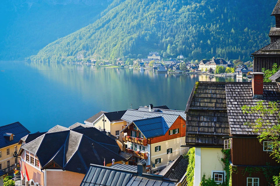A hillside view of a lakeside village with various houses, a calm lake, and forested mountains in the background.