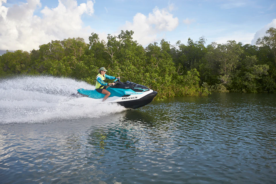 Person riding a teal and black jet ski on a lake, with green trees and a partly cloudy sky in the background.