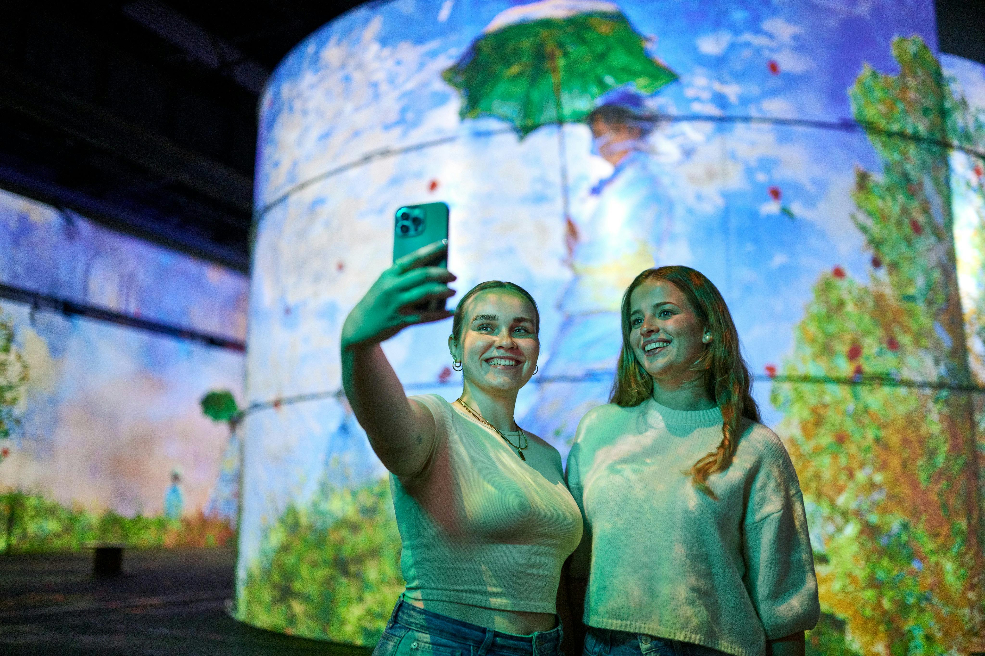 Δύο γυναίκες χαμογελούν για μια selfie μπροστά από μια μεγάλη οθόνη που εμφανίζει μια ζωγραφισμένη φιγούρα που κρατά μια πράσινη ομπρέλα.