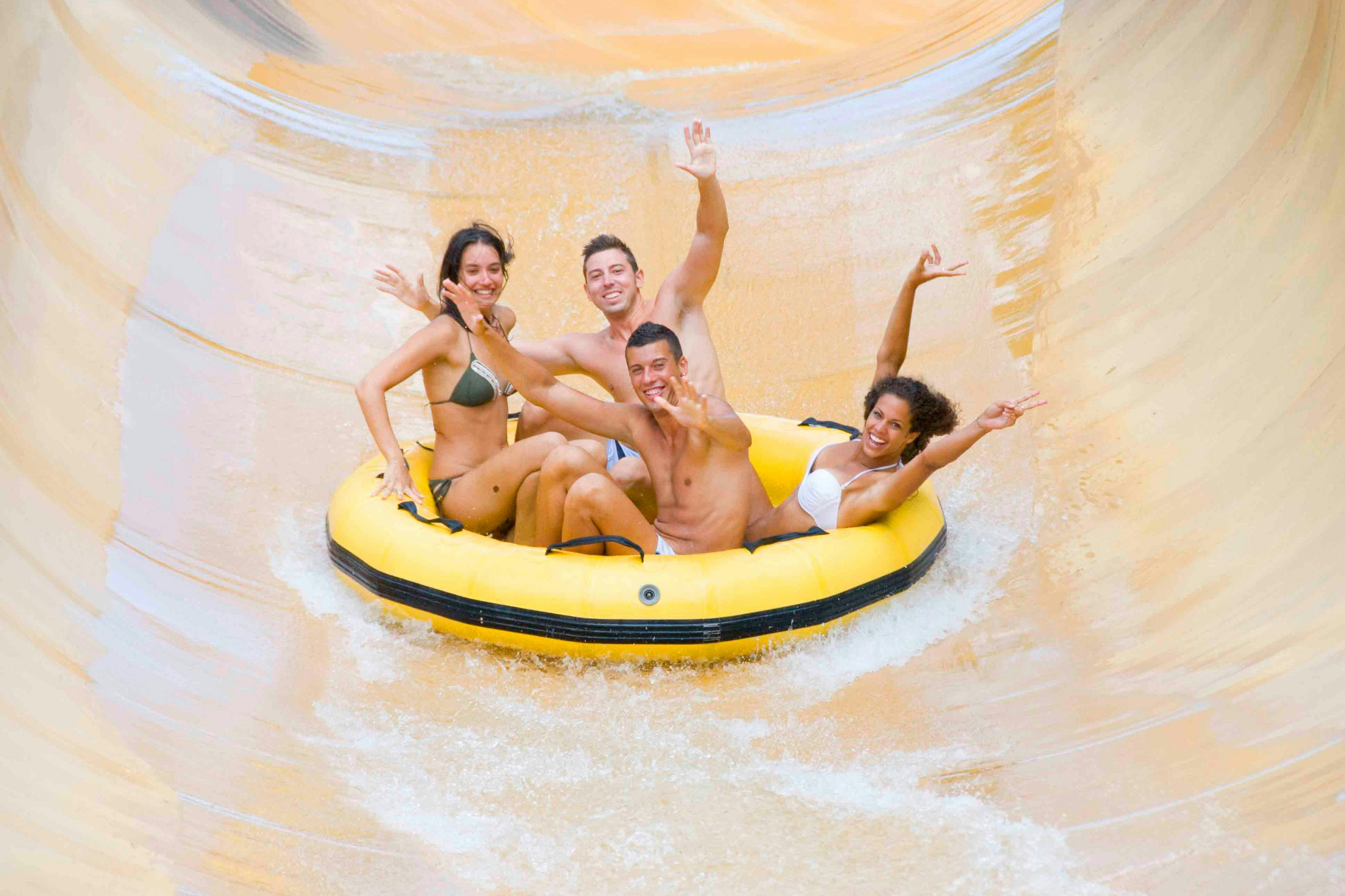 Vier mensen rijden op een geel opblaasbaar vlot van een waterglijbaan, glimlachen en steken opgewonden hun armen omhoog.