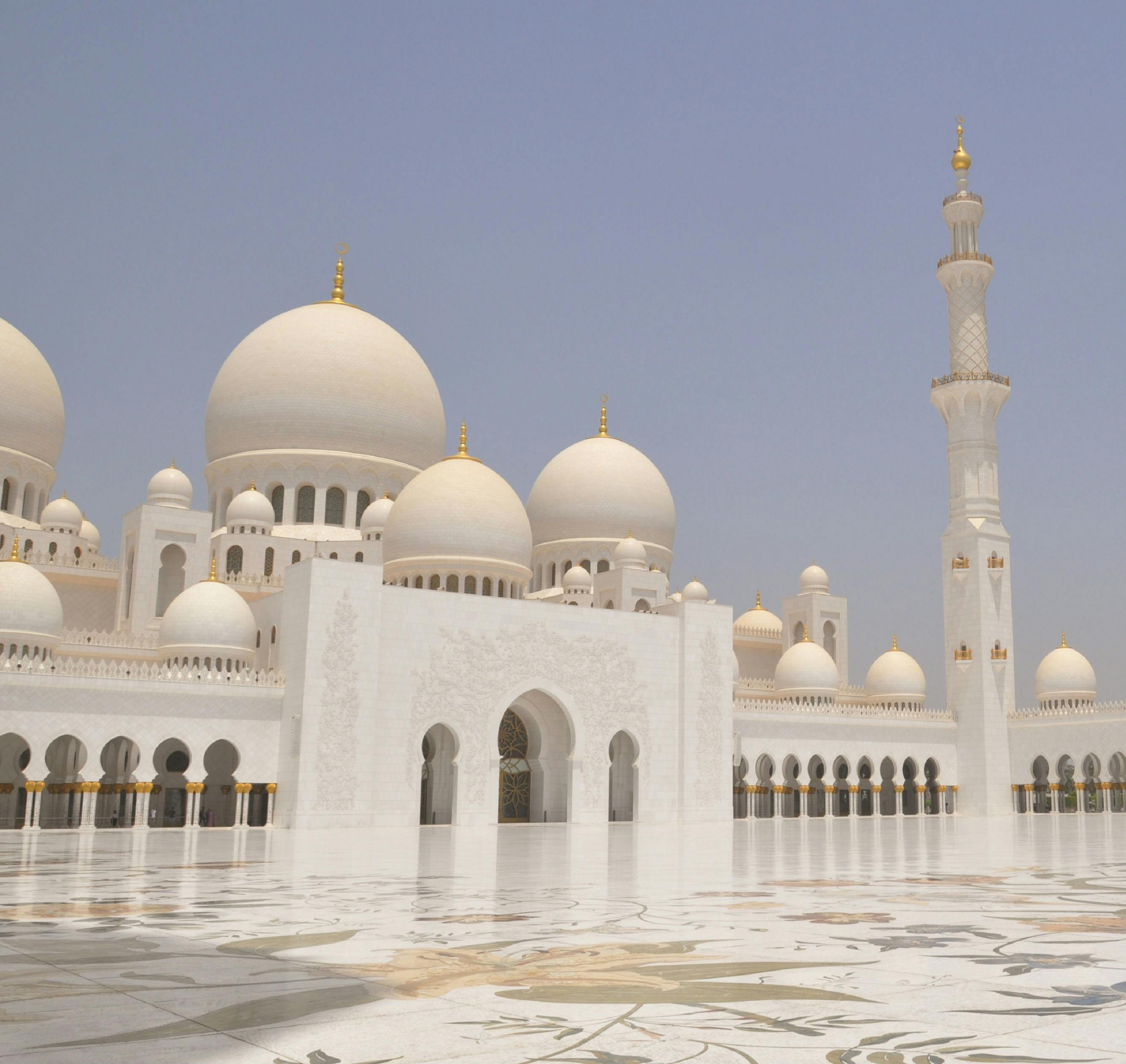 White mosque with large domes and minaret, intricate carvings, and spacious courtyard under clear blue sky.