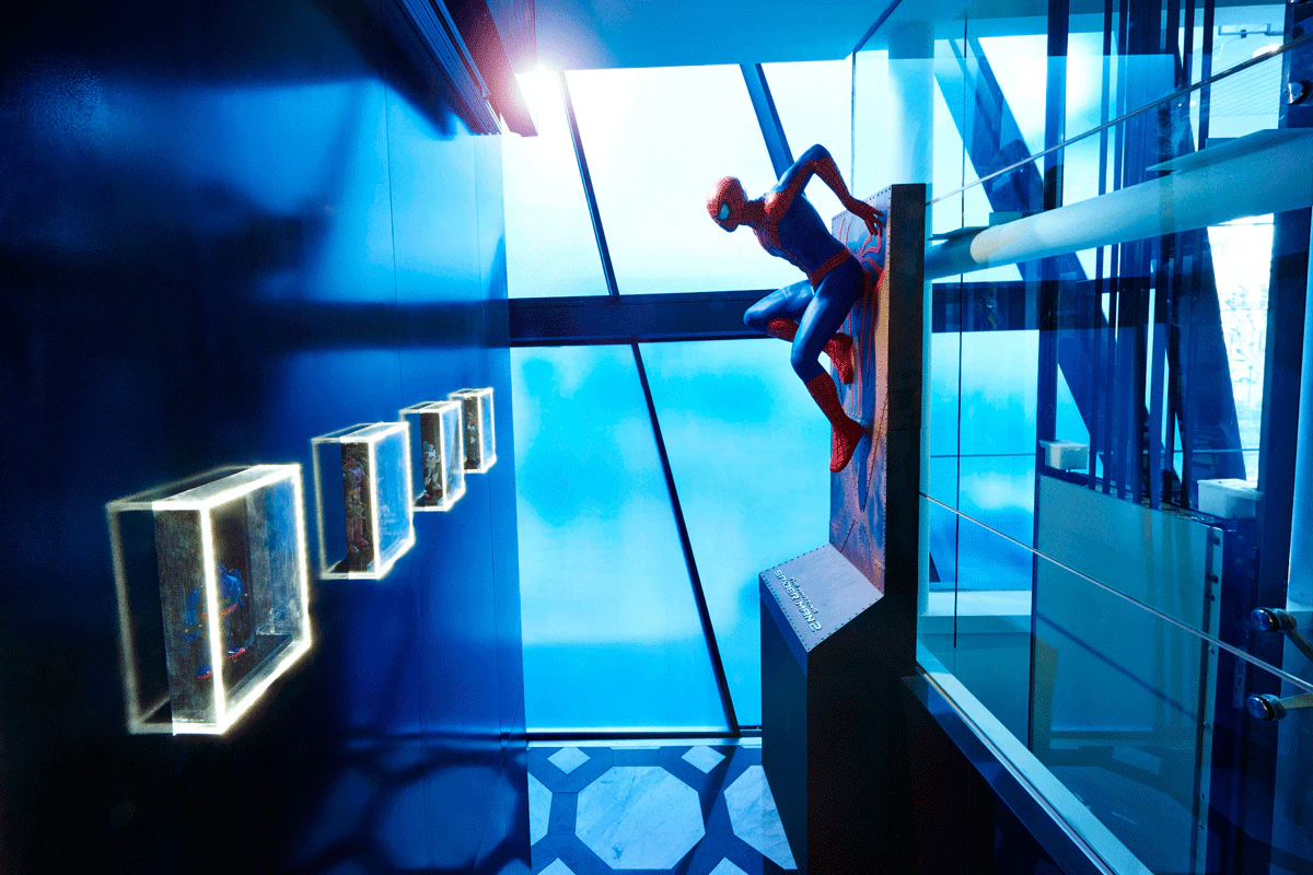 A Spider-Man statue mounted on a wall in a modern, blue-lit exhibition space with illuminated display cases on the left.