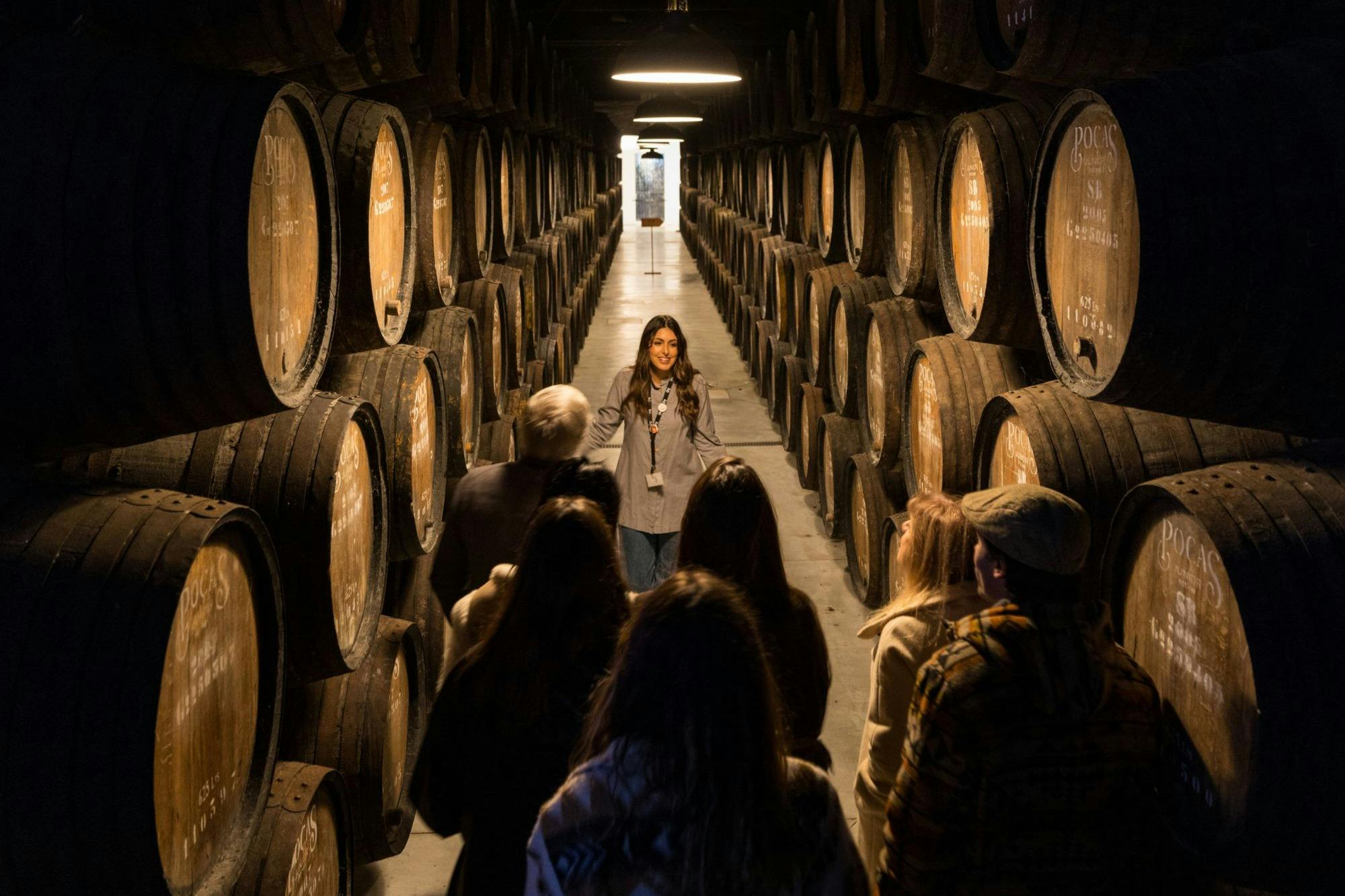 A group of people listening to a guide inside a wooden barrel storage room, with barrels lining both sides of the aisle.