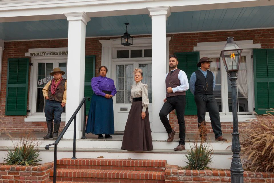 Five people dressed in historical attire stand on the steps of a brick building with green shutters and white columns.