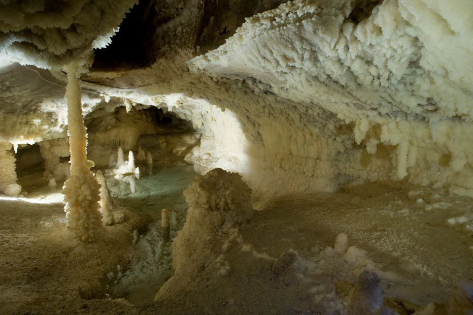 A cave interior with stalactites and stalagmites illuminated, showing natural rock formations and a small pool of water.