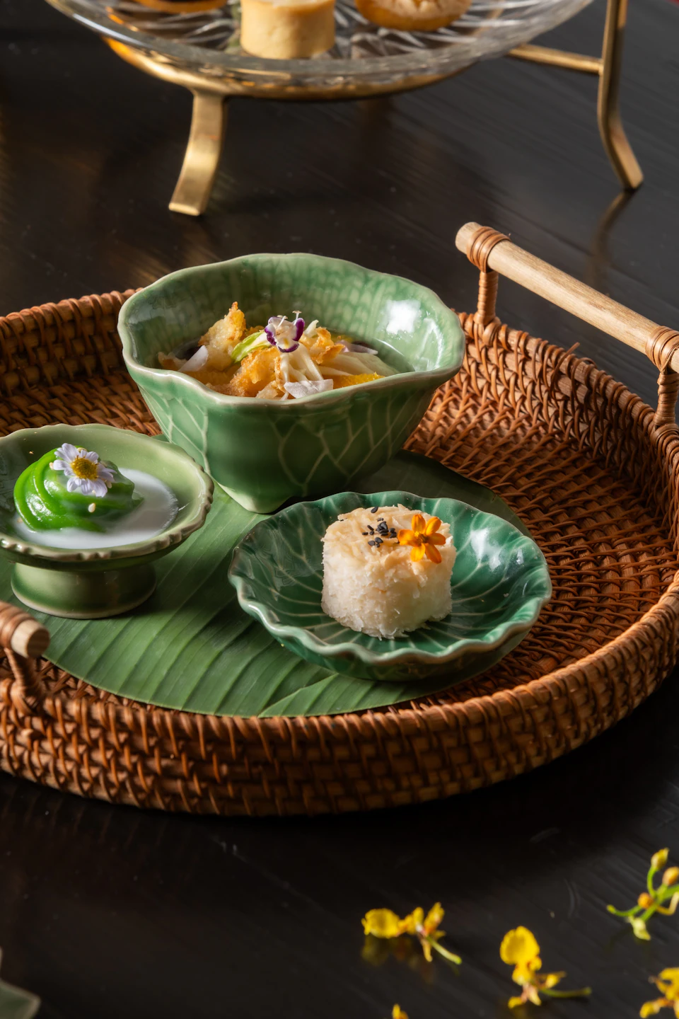 A woven tray holding three green ceramic dishes with decorative food, placed on a banana leaf, on a dark surface.
