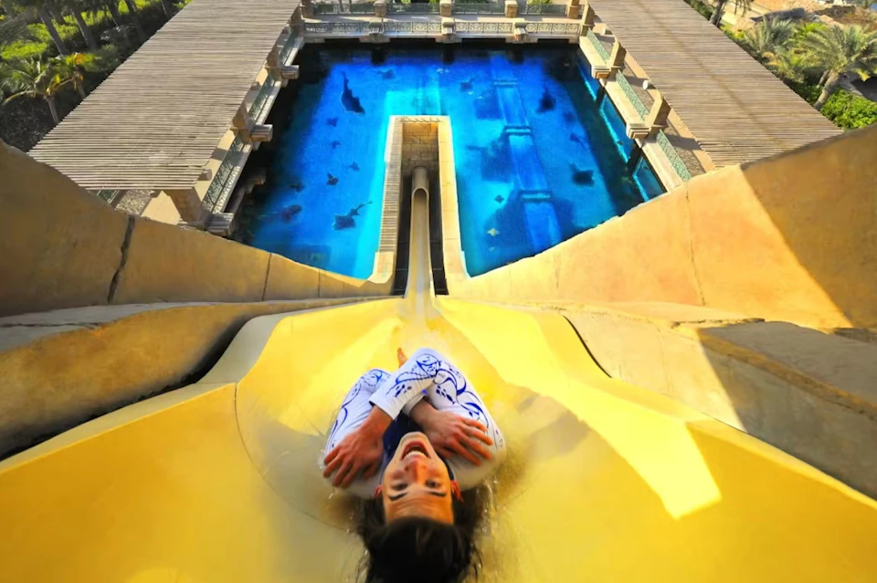 Person sliding down a yellow water slide into a pool with visible sharks, surrounded by a structure with wooden decks.