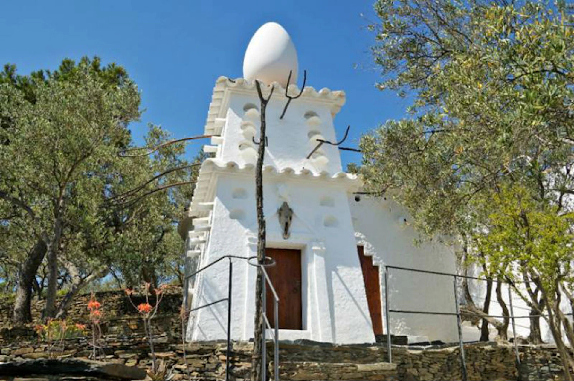White building with an unusual egg-shaped sculpture on the roof, surrounded by trees and a clear blue sky.