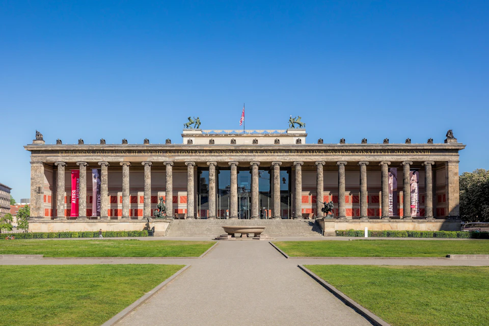 A neoclassical building with a colonnade, statues on the roof, and a flag, surrounded by greenery under a blue sky.