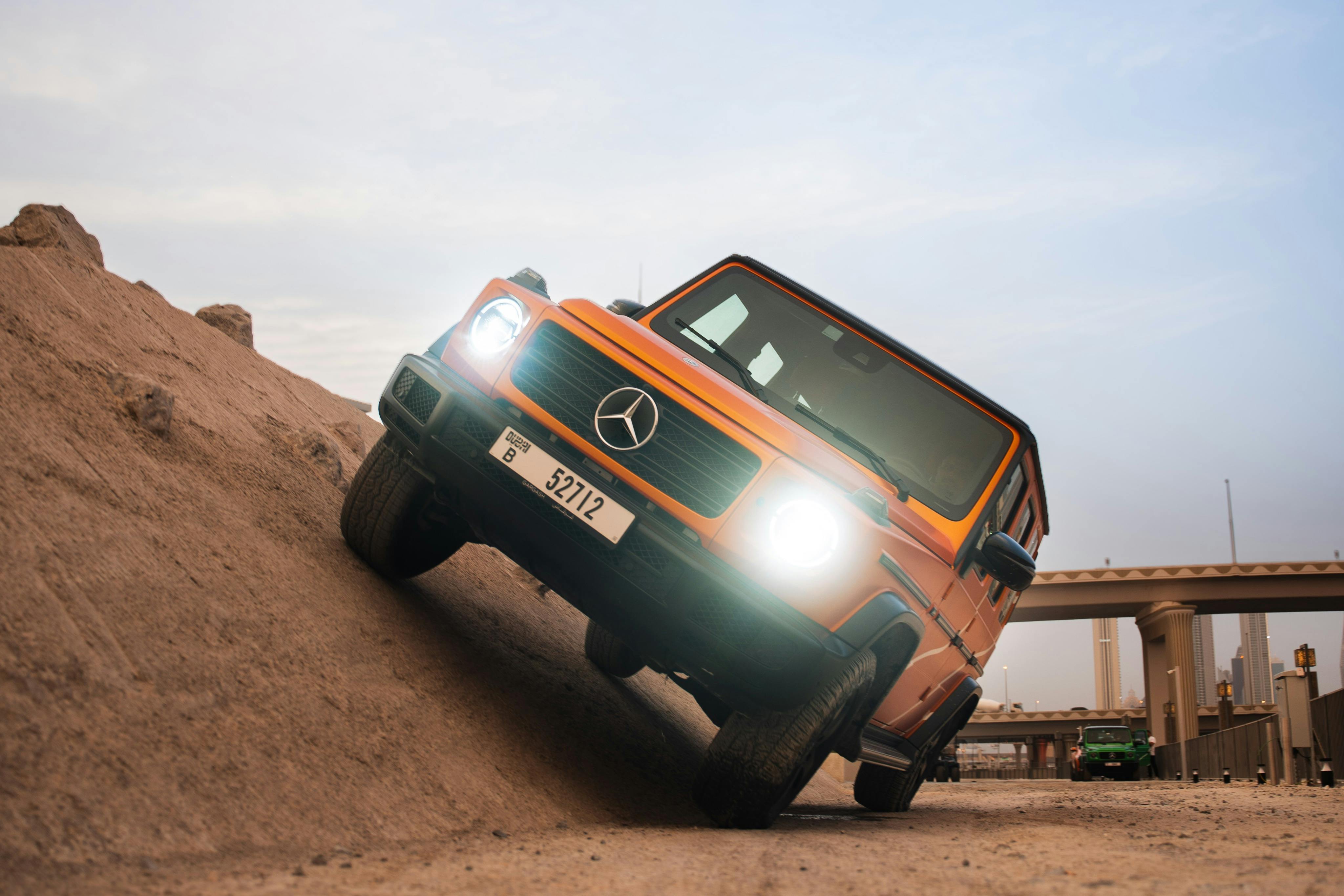A bright orange Mercedes G-Class SUV ascending a steep dirt slope, with headlights on and slightly tilted to one side.