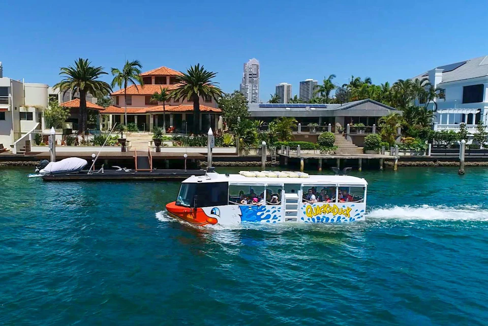 A boat painted like a duck with "Quack'R'Duck" on the side floats on a river near waterside houses and palm trees under a clear blue sky.